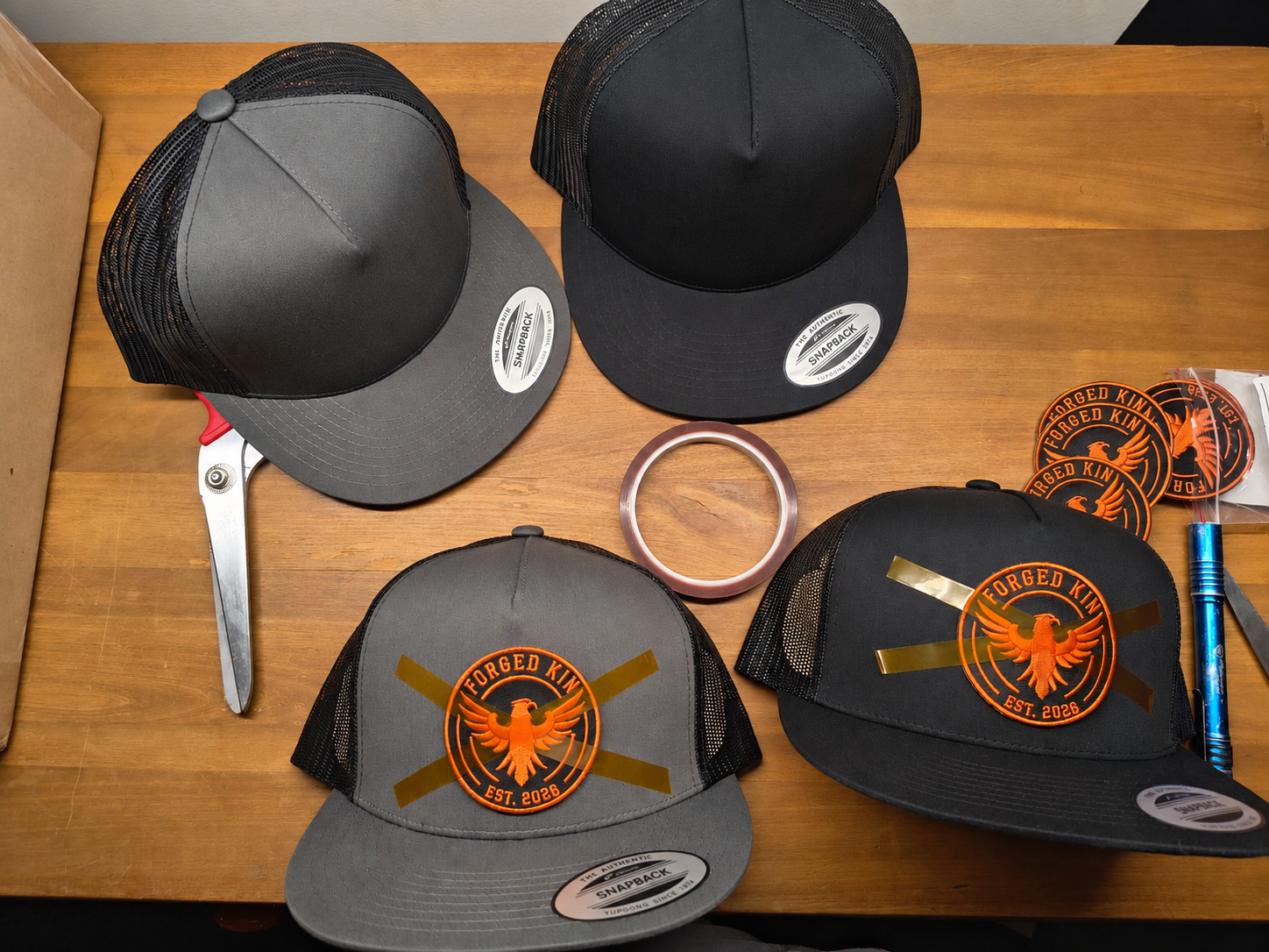 FORGED KIN hat development: blank caps, eagle patches, tape and scissors on a wooden table