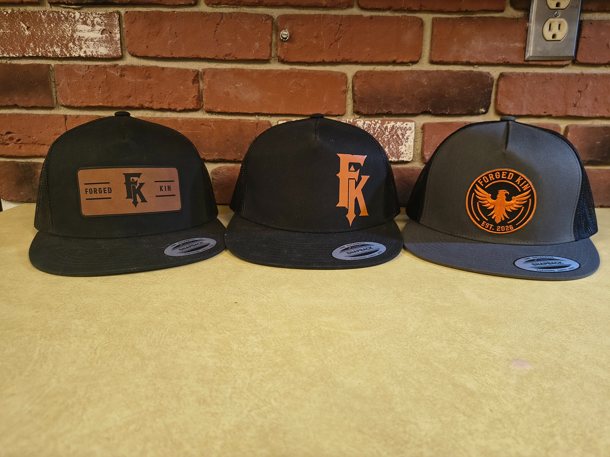 Three FORGED KIN hats with a faux leather patch, FK monogram and circular eagle patch against a brick wall